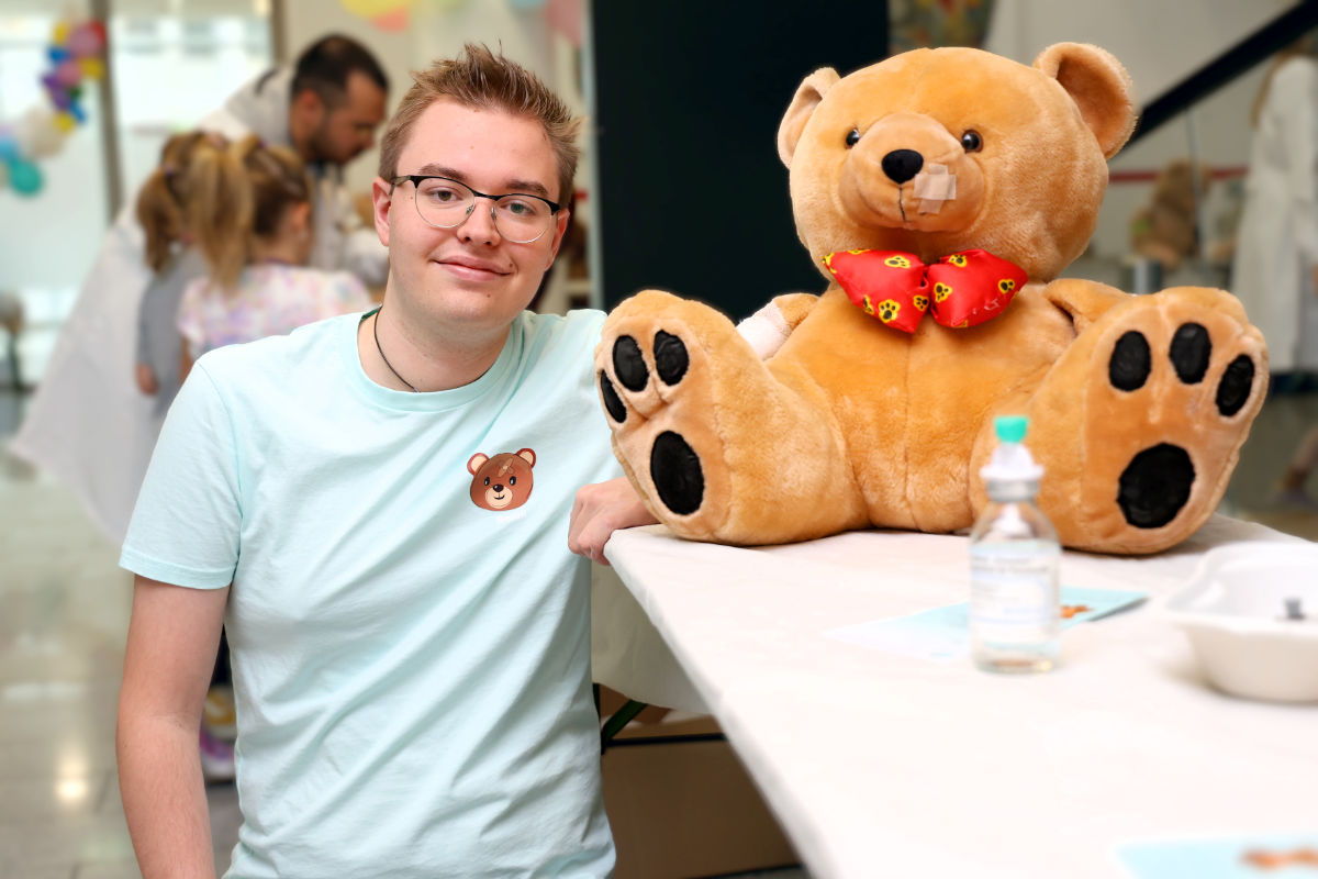 Medizinstudent Benedikt Papst lehnt lächelnd an einem Tisch neben einem großen Teddy mit roter Schleife – Szene aus dem Teddybärkrankenhaus Innsbruck.