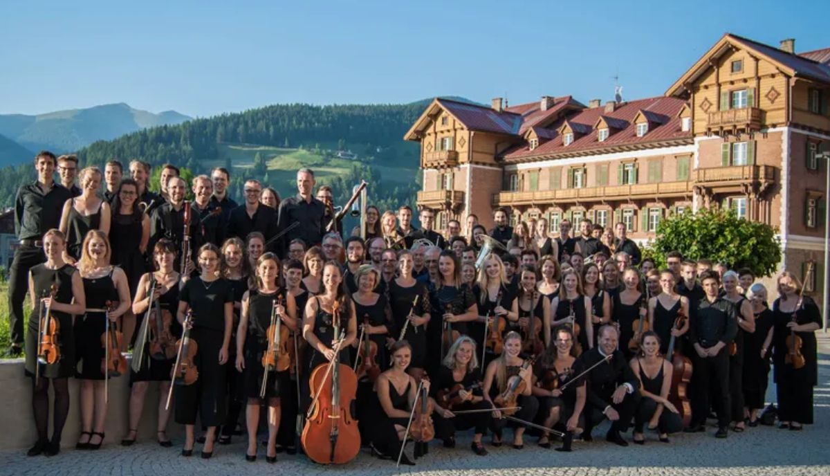 Die Mitglieder des Universitätsorchesters stehen mit ihren Instrumenten vor einem Gebäude