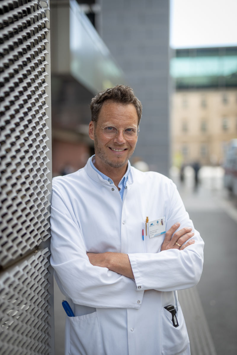 Gregor Brössner, im weißen Kittel, lehnt lächelnd und mit verschränkten Händen an einer Mauer im Klinikareal.